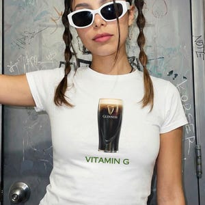 May include: White t-shirt featuring a graphic of a pint of Guinness stout with the text "VITAMIN G" in green. The shirt is a crew neck style.