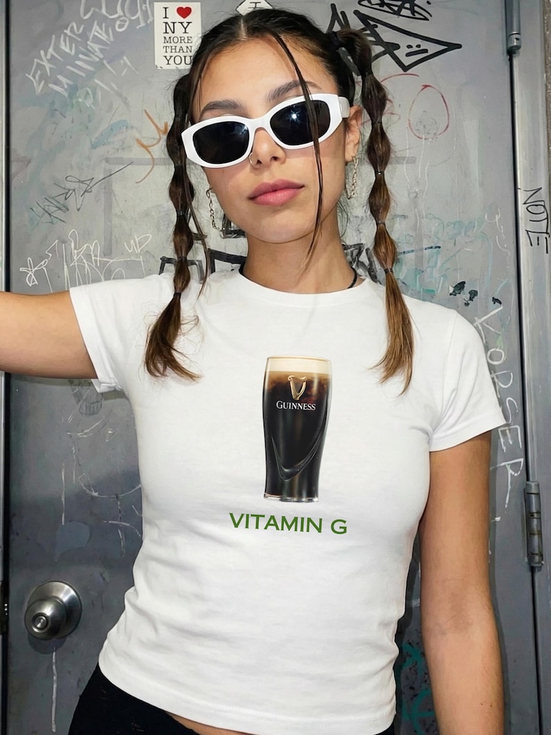 May include: White t-shirt featuring a graphic of a pint of Guinness stout with the text "VITAMIN G" in green. The shirt is a crew neck style.