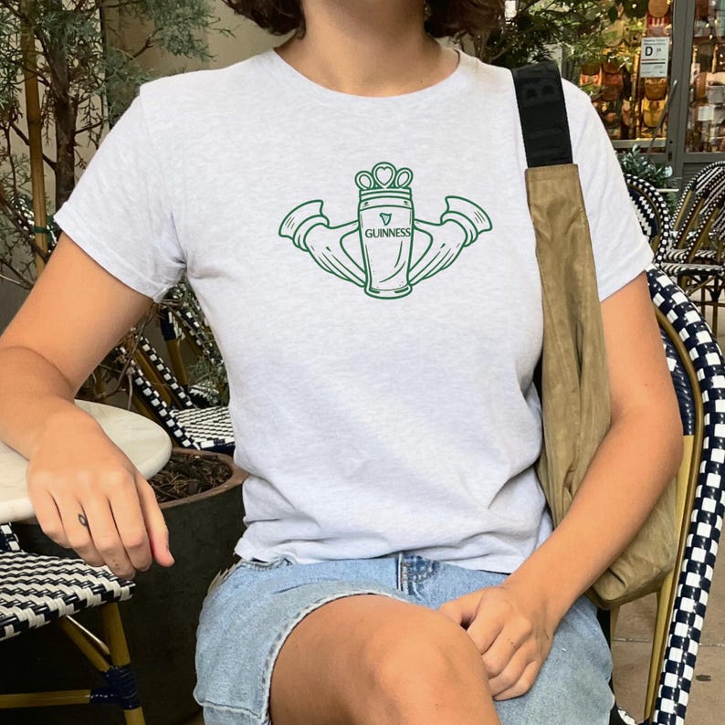 May include: A light grey t-shirt with a green graphic of a Guinness pint glass, crown, and hands. The word "Guinness" is printed on the glass. The shirt is short-sleeved.
