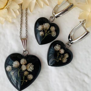 May include: A black heart-shaped pendant necklace and matching earrings with delicate white flowers pressed inside the resin. The jewelry is on a white background.