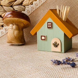 Könnte beinhalten: Ein dekorativer Zahnstocherhalter in Form eines Holzhauses mit hellgrünem Körper und orangefarbenem Dach. Das Haus hat eine kleine Tür mit einem bienenförmigen Griff und zwei kleine Fenster. Links ist ein Keramikpilz.