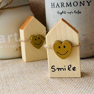 May include: Two small, wooden house-shaped ornaments with gold heart accents and the word "Smile" written on one. The ornaments are tied with twine and sit on a textured surface, with a candle and a concrete pot in the background.