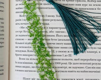 May include: A handmade bookmark with a green, fern-like design and a teal tassel. The bookmark is placed on a book, adding a touch of nature to reading. The bookmark is a unique accessory for book lovers.