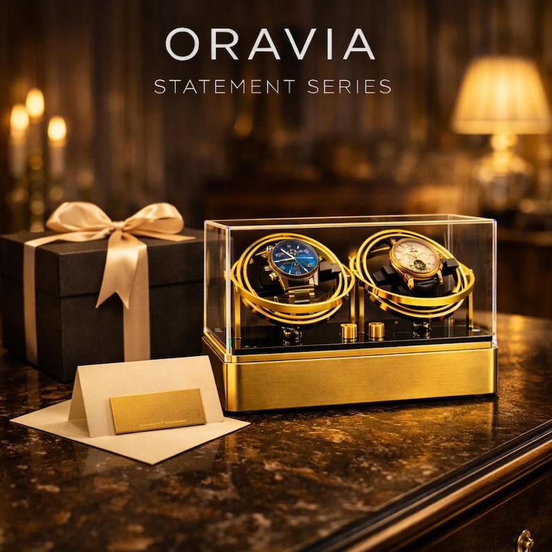 May include: A gold and clear watch display case holding two watches, one with a blue face and the other with a white face. The case has the text "ORAVIA STATEMENT SERIES". A black gift box with a gold ribbon and a gold card are also visible.