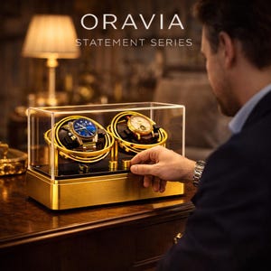 May include: A gold and black watch winder with two watch holders, each holding a wristwatch. The winder is enclosed in a clear case and sits on a wooden surface. The text "ORAVIA STATEMENT SERIES" is displayed above the winder.