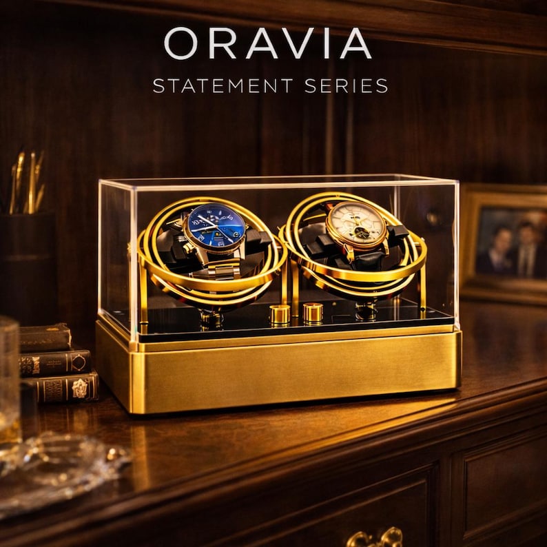 May include: A gold and black watch winder with two compartments, each holding a wristwatch. The watches have blue and white faces. The winder is in a clear acrylic case with the text "ORAVIA STATEMENT SERIES" at the top.