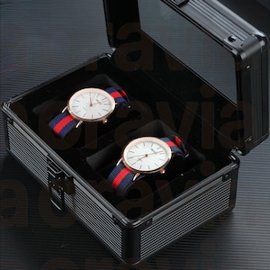 May include: Two gold-coloured wristwatches with white faces and red, white, and blue striped straps are displayed in an open black case. The case has a textured exterior and a black velvet interior.