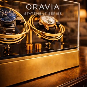 May include: A gold-coloured watch display case with two watches inside. The case has a clear glass top and the text "ORAVIA STATEMENT SERIES" is visible. The watches have gold and silver tones with black watch straps.