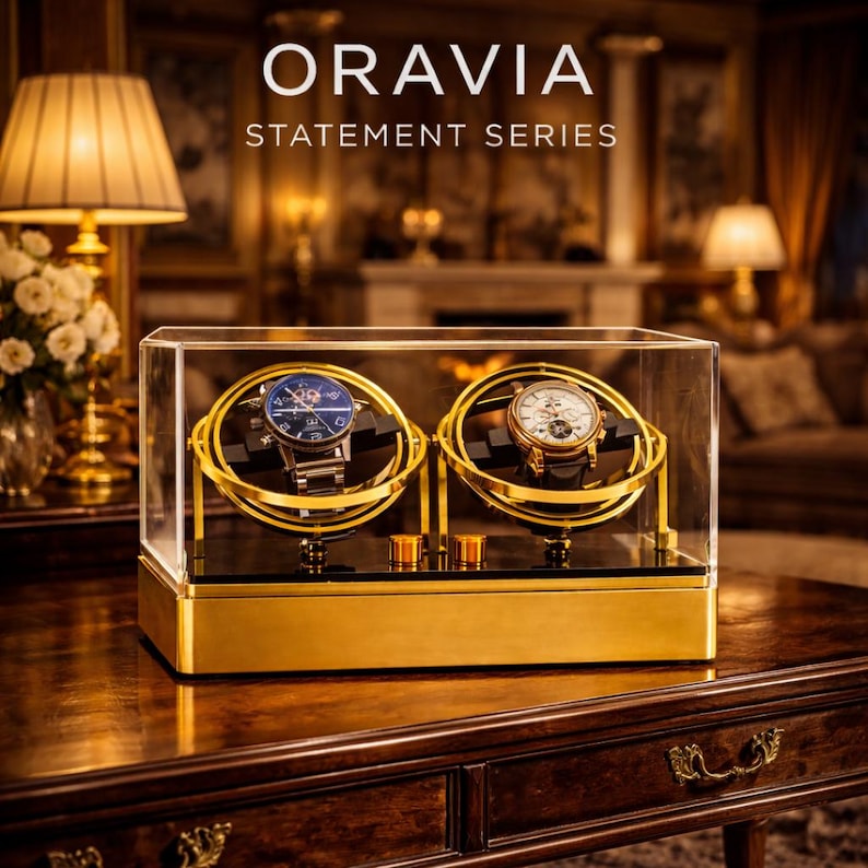 May include: A gold and clear watch display case with two watches. The case has the text "ORAVIA STATEMENT SERIES". The watches are on black holders, and the case sits on a dark wood table.