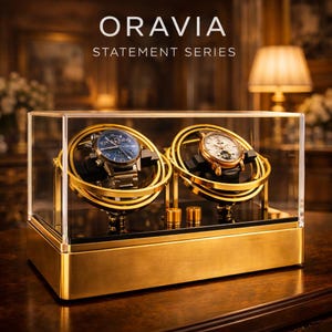 May include: A gold-coloured watch display case with two watches inside. The case has a clear glass top and sides, with the text "ORAVIA STATEMENT SERIES" at the top. The watches have blue and white faces, and gold-coloured bands. The case sits on a wooden surface.