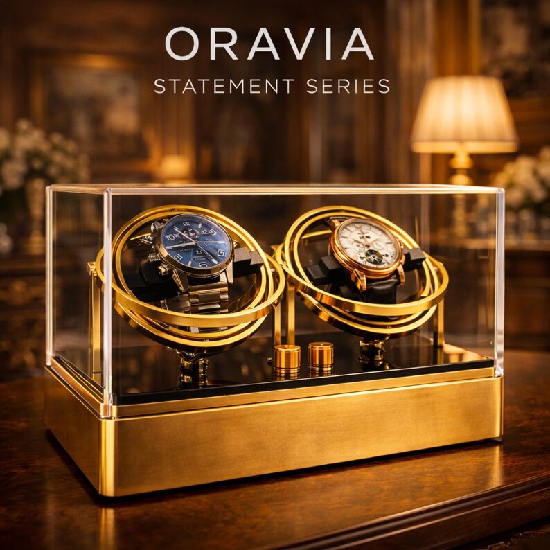 May include: A gold-coloured watch display case with two watches inside. The case has a clear glass top and sides, with the text "ORAVIA STATEMENT SERIES" at the top. The watches have blue and white faces, and gold-coloured bands. The case sits on a wooden surface.