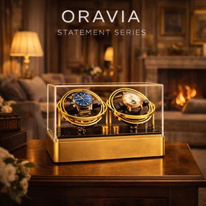 May include: A gold watch display case with two watches inside. The watches have blue and white faces. The case is labelled "ORAVIA STATEMENT SERIES". The case sits on a wooden table in a room with a fireplace and lamps.