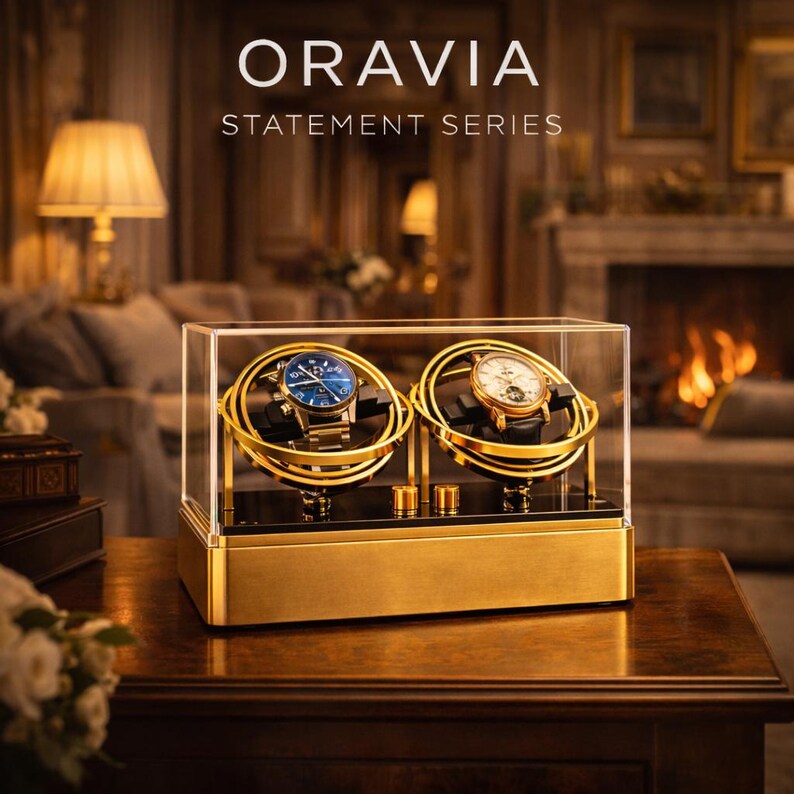 May include: A gold watch display case with two watches inside. The watches have blue and white faces. The case is labelled "ORAVIA STATEMENT SERIES". The case sits on a wooden table in a room with a fireplace and lamps.