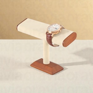 May include: A watch display stand with a cream-colored padded top and a brown leather watch. The stand has a wooden base and accents. The watch has a brown leather band and a gold-colored face.