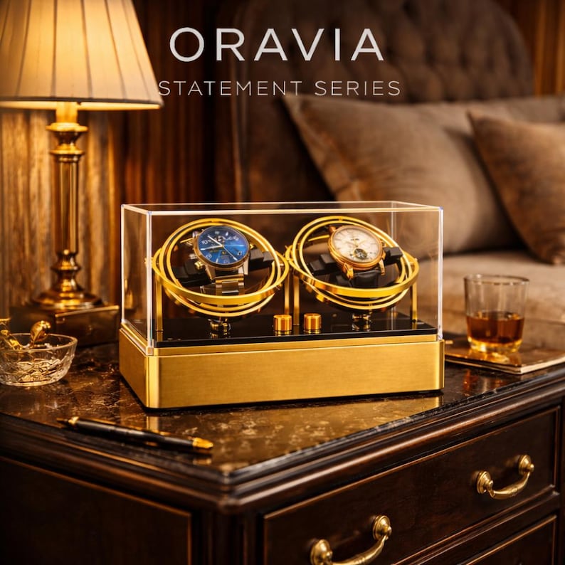 May include: A gold watch display case with two watches inside. The watches have blue and white faces. The case is labelled "ORAVIA STATEMENT SERIES". The case sits on a dark wood surface with a lamp and a glass of amber liquid in the background.