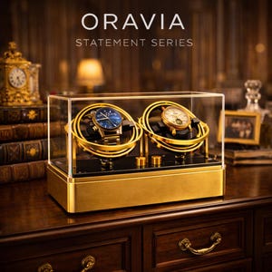 May include: A gold and black watch winder with two watches inside a clear glass case. The watches have blue and white faces. The winder is on a wooden surface. The text "ORAVIA STATEMENT SERIES" is at the top.