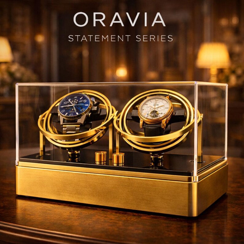 May include: A gold-coloured watch winder with two watch holders, enclosed in a clear case. The winder has the text "ORAVIA STATEMENT SERIES" on top. Two watches with blue and white faces are displayed.