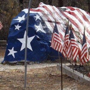 May include: A large rock painted with an American flag design. Several smaller American flags are displayed in front of the rock.