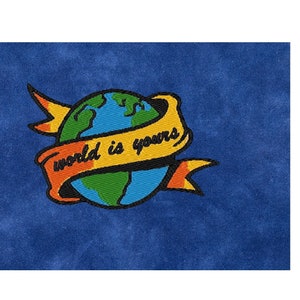 May include: Embroidered patch with a blue globe surrounded by a yellow ribbon with the text "world is yours" in black lettering. The ribbon is outlined in black and has red and orange accents.