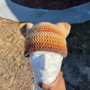 May include: A crochet hat in brown, tan, and orange, with ear details. The hat is displayed on a white mannequin head. The hat's colours and design are highlighted. A cozy accessory, perfect for cooler weather.