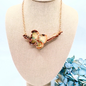 May include: A handcrafted necklace featuring a unique floral pendant. The pendant showcases a multi-colored flower design with shades of brown, green, and cream, attached to a gold-colored chain. The necklace is displayed on a beige mannequin.