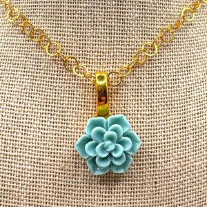 May include: A gold-colored chain necklace with a pendant featuring a light blue succulent flower. The pendant is attached to a gold-colored bail. The necklace is displayed on a neutral-colored fabric.
