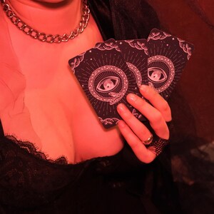 May include: Two tarot cards with a dark background and a pink eye and snake design are held in a hand. The cards have a circular design with radiating lines and a crescent moon and stars. A silver chain necklace is visible.
