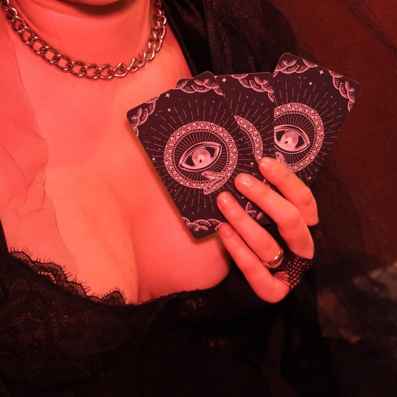 May include: Two tarot cards with a dark background and a pink eye and snake design are held in a hand. The cards have a circular design with radiating lines and a crescent moon and stars. A silver chain necklace is visible.