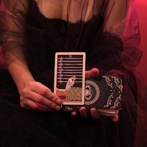 May include: A tarot card from the Nine of Swords suit, featuring a person in a white robe sitting on a bed with nine swords above. The card is held with a deck of cards with a celestial design.