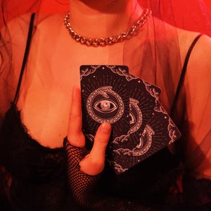 May include: A pair of black tarot cards featuring an eye within a serpent design, held in front of a black top. The cards have a celestial pattern with stars and clouds. A gold chain necklace is visible.