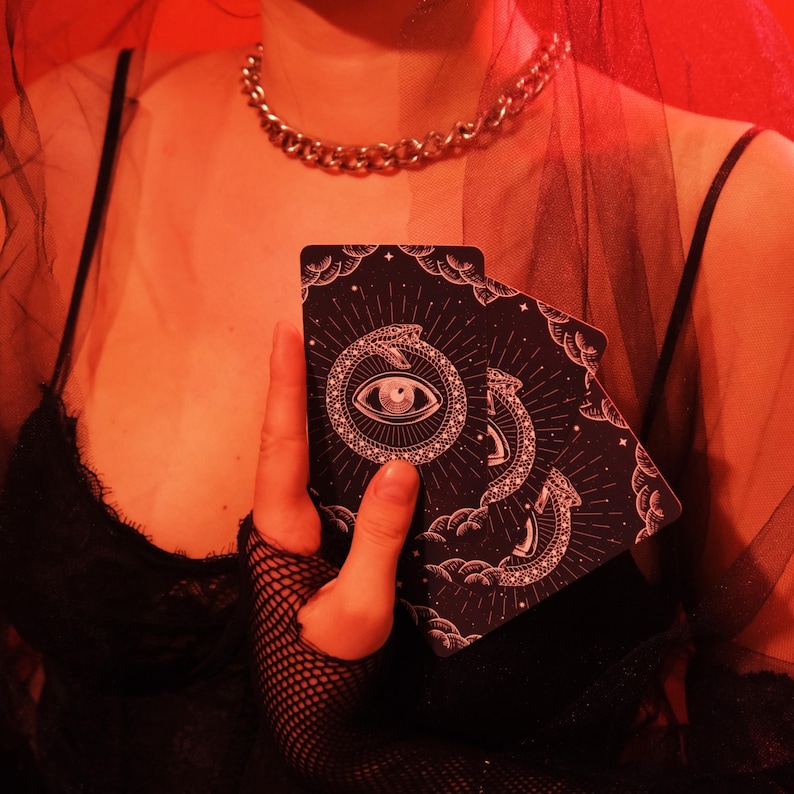 May include: A pair of black tarot cards featuring an eye within a serpent design, held in front of a black top. The cards have a celestial pattern with stars and clouds. A gold chain necklace is visible.