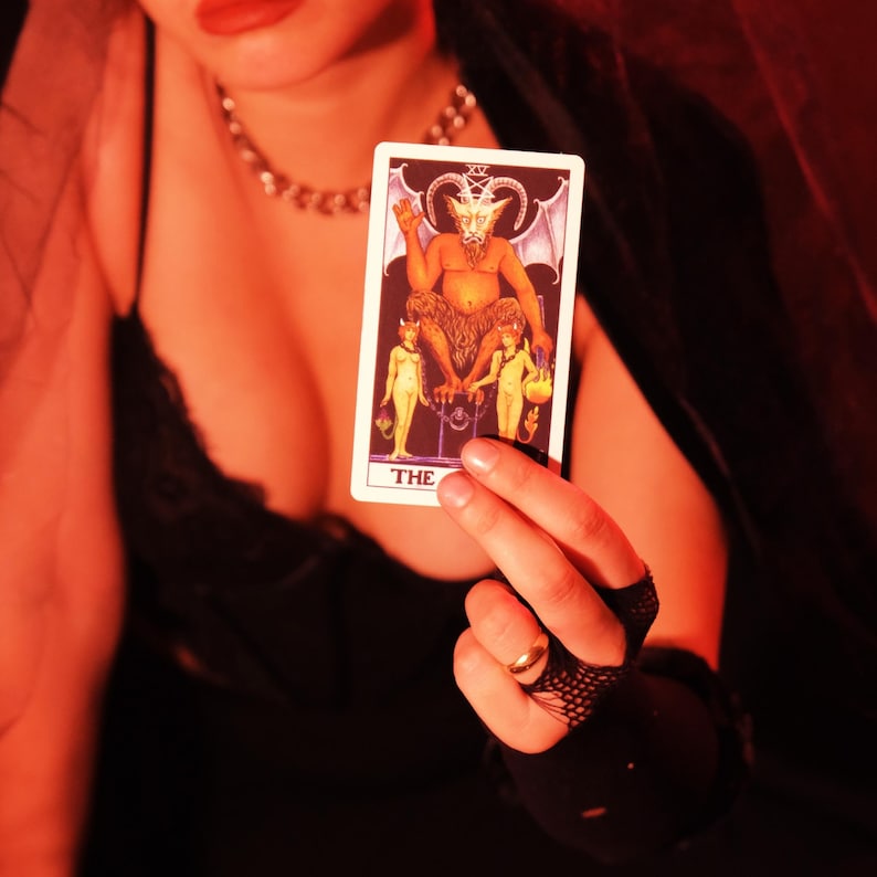 May include: A tarot card featuring a horned figure with wings, seated between two smaller figures. The card has the word "THE" at the bottom. The card is held in a hand with a gold ring, against a dark background.