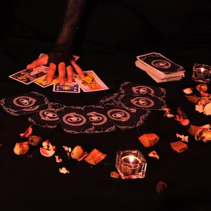 May include: A tarot card reading setup with a hand wearing a black fishnet glove hovering over a spread of cards. The cards feature celestial and symbolic imagery. A stack of cards, lit candles, and dried flowers are also present.