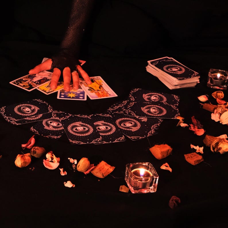 May include: A tarot card reading setup with a hand wearing a black fishnet glove hovering over a spread of cards. The cards feature celestial and symbolic imagery. A stack of cards, lit candles, and dried flowers are also present.