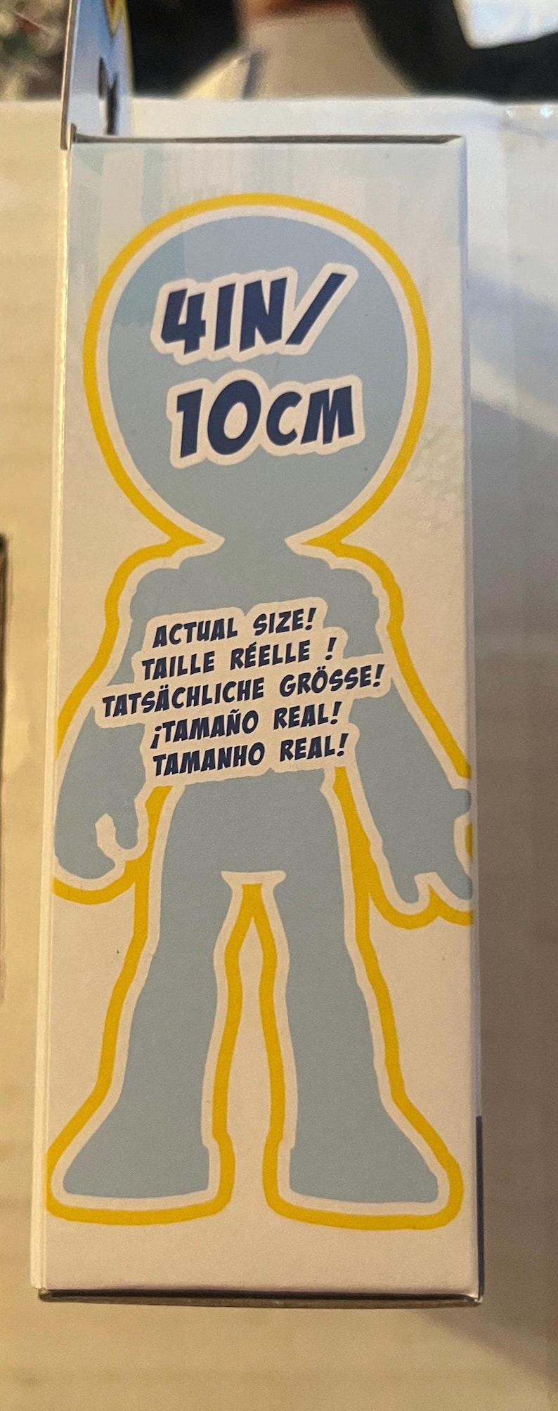 May include: A product box with a light blue silhouette of a person, outlined in yellow. The text on the box indicates the actual size is 4 inches / 10 cm. Additional text includes "Actual Size!" in multiple languages.
