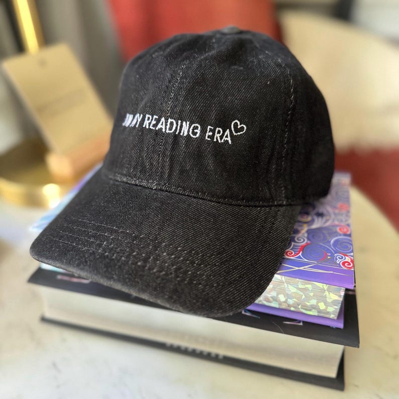 以下が含まれることがあります： 「MY READING ERA」の刺繍と小さなハートが付いた黒いデニムの野球帽。本の上に置かれています。つばはカーブしており、クラウンは構造化されています。