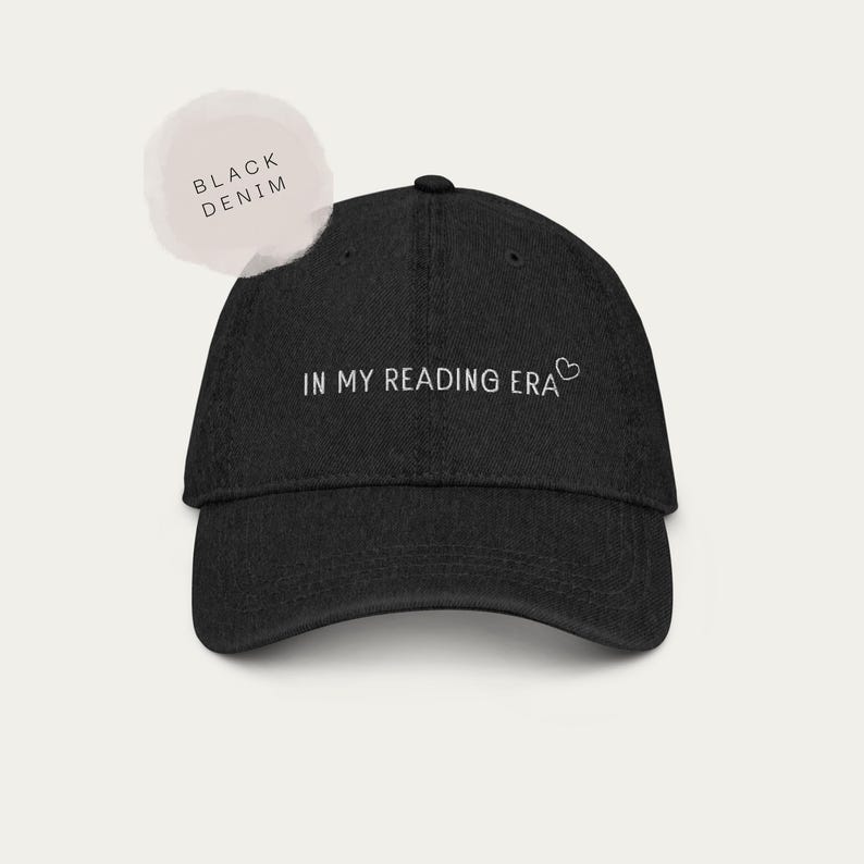 以下が含まれることがあります： 黒色のデニムベースボールキャップ。白い文字で「IN MY READING ERA」と小さなハートのシンボルがプリントされています。キャップはカーブしたつばと構造化されたクラウンを備えています。明るい色の円形グラフィックには「BLACK DENIM」と書かれています。