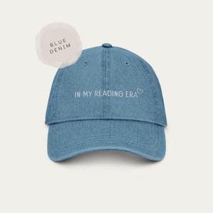 以下が含まれることがあります： 「IN MY READING ERA」の白い文字と小さなハートのグラフィックが入った、青いデニムの野球帽。つばはカーブしており、上部にボタンが付いています。「BLUE DENIM」と書かれたラベルがあります。