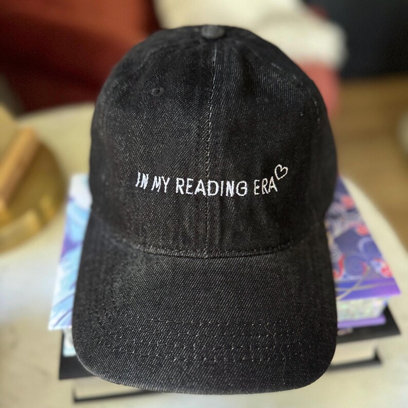 以下が含まれることがあります： 白い文字で「IN MY READING ERA」と刺繍された黒いデニムの野球帽。小さなハートのシンボルが付いています。帽子はカーブしたつばと上部にボタンが付いています。