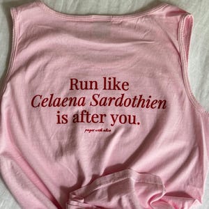 Può includere: Canotta rosa senza maniche con il testo "Run like Celaena Sardothien is after you." in lettere rosse. Il testo "pages with alice" è anche stampato in lettere rosse.