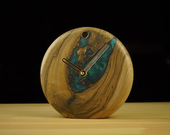 May include: A round wooden clock with a blue and green resin inlay. The clock has gold-colored hands and a simple design.