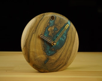 May include: A round wooden clock with a teal resin inlay. The clock face features gold-colored hands and a small gold accent at the top. The wood grain is visible, and the clock sits on a wooden surface.