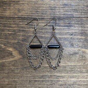 May include: A pair of silver-toned metal dangle earrings featuring a horizontal black cylindrical bead suspended within a teardrop-shaped frame. The design includes multiple delicate silver chains draped beneath the central bead, attached to a standard hook.