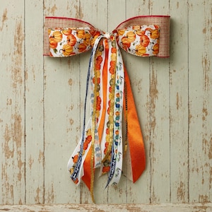 May include: A decorative bow made of multiple ribbons featuring a pumpkin pattern, solid orange satin, and white ribbon with the word home printed in black. The bow includes a tan plaid border and long, cascading ribbon tails of varying widths.