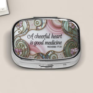 May include: A silver pill box with a decorative top featuring the quote "A cheerful heart is good medicine" from Proverbs 17:22. The design includes pink, green, and gold accents with floral and swirl motifs.