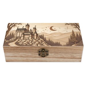 May include: Wooden box with a detailed carved design of a castle on a hill, trees, a crescent moon, and stars. The box has a metal clasp and is made of light-colored wood. The artwork is in shades of brown and beige.