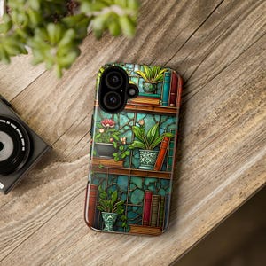 May include: A smartphone case featuring a stained-glass design of a bookshelf with potted plants and books. The case has a glossy finish and a detailed, colorful illustration with shades of green, brown, and red. The case is resting on a wooden surface.