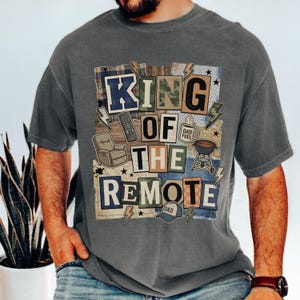 Puede incluir: Camiseta gris con un gráfico colorido que dice "KING OF THE REMOTE". El diseño incluye un mando a distancia, un sillón reclinable, una parrilla y rayos. La camiseta tiene un corte holgado.