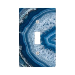 May include: A decorative light switch cover featuring a blue and white agate stone pattern. The cover has a single toggle switch opening and two silver screws. The design showcases concentric bands of blue and white, creating a natural, layered effect.