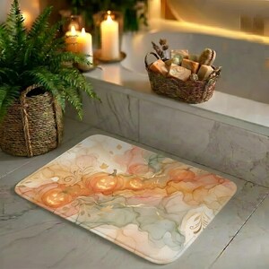 Op de afbeelding: Een rechthoekige badmat met een zachte aquarelprint van oranje pompoenen, abstracte crème- en saliegroene wolken en delicate goudkleurige lijntekeningen op een lichte, neutrale achtergrond.